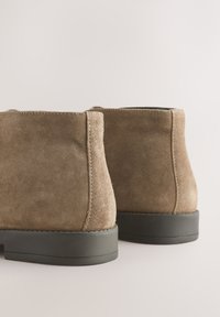 Hnedé členkové čižmy z brúseného semišu s textúrovaným povrchom, zaoblenou špičkou a nízkou čiernou gumovou podrážkou, so švami viditeľnými na zadnej strane.