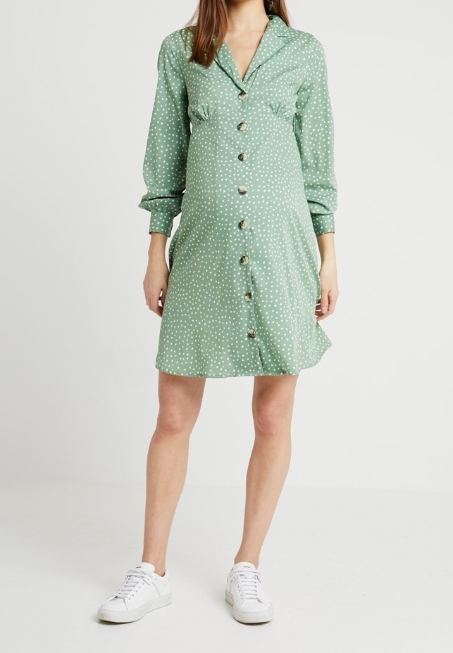 Femme portant une robe verte à manches longues boutonnée avec des pois blancs et des baskets blanches, debout devant un fond uni.