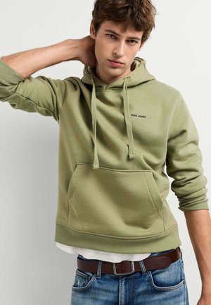 Giovane uomo con capelli castani che indossa una felpa verde chiaro con cappuccio e jeans blu, posando con una mano dietro il collo su uno sfondo semplice.