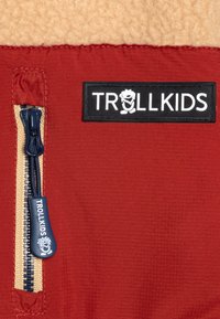 Punane jakk beežist fliisist ülikonna peal. Omab külgvetti, millel on tumesinine tõmblukk ja must "TROLLKIDS" logoplekk. Tekstuurne kangas.