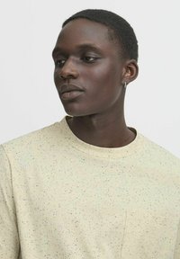 Lys beige t-skjorte med flekkete mønster, rund hals og brystlomme. Myk tekstur med korte ermer, vist brukt av en person.