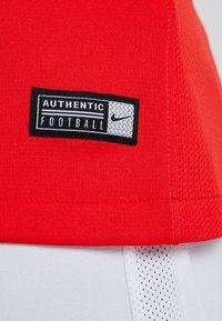 Tejido deportivo rojo con una etiqueta negra y blanca de Nike "Fútbol Auténtico" cosida cerca de una franja de malla blanca.