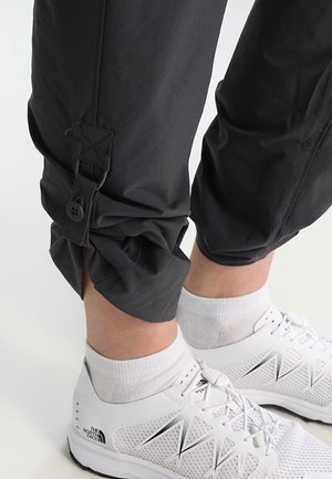 Pantalons noirs légers avec des poignets froncés, équipés de boutons et de fermetures à pression. Associés à des chaussures de sport blanches et des chaussettes blanches.