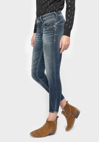 Jeans skinny de mezclilla azul con áreas desgastadas y pequeños desgarros. Tienen una cintura de altura media y se combinan con botines marrones.