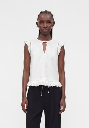 Femme aux cheveux tressés portant un chemisier blanc sans manches avec des bords volantés et un pantalon noir taille haute sur un fond uni.