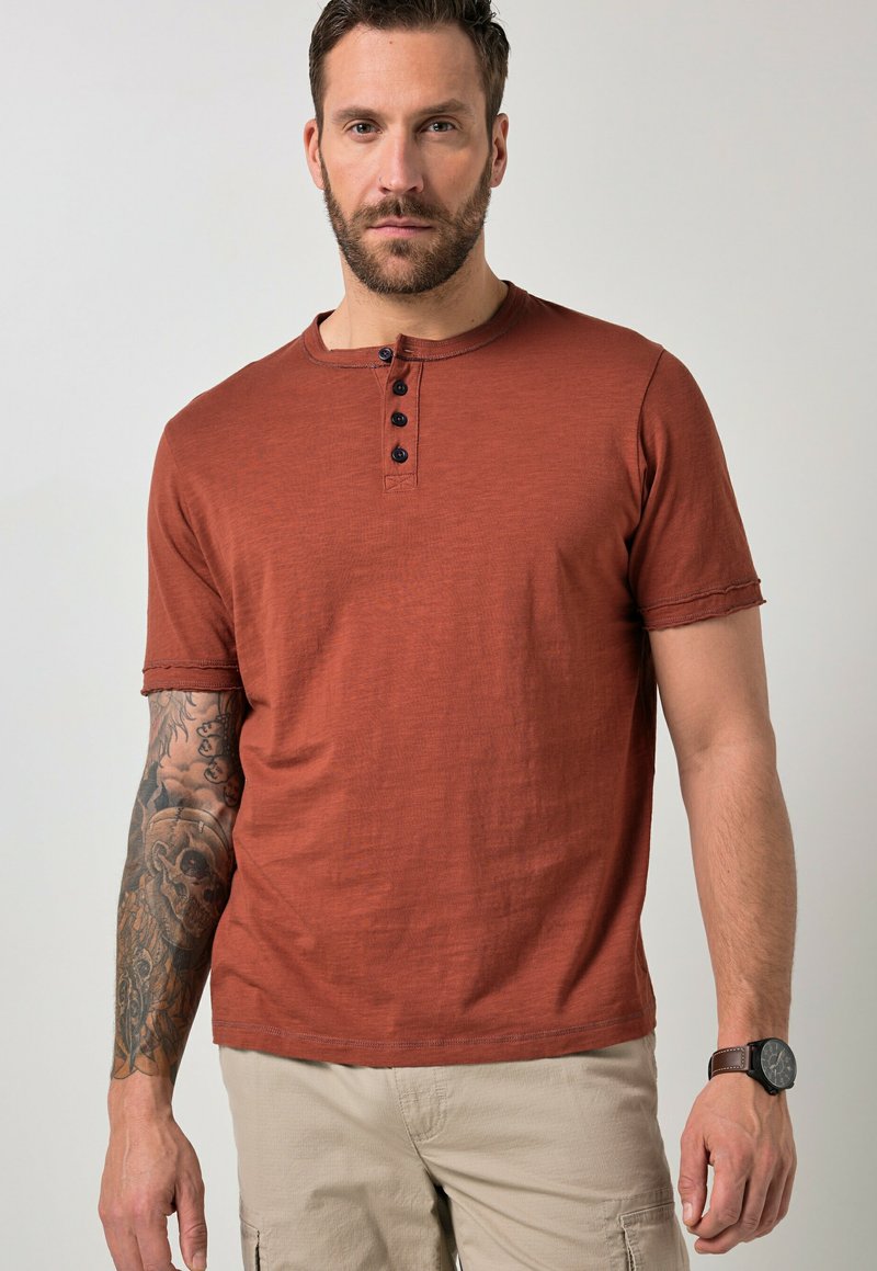 Hombre vistiendo una camisa henley de manga corta color óxido, pantalones beige, reloj en la muñeca izquierda y tatuaje de manga en el brazo derecho, de pie contra un fondo liso.