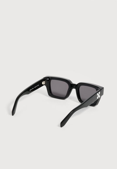 Lunettes de soleil rectangulaires à monture épaisse noire avec verres foncés, branches pliées et logo croix blanc sur la branche droite, sur fond blanc.