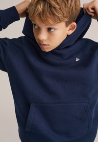 Tumesinine kapuutsviit, mis on valmistatud pehmest puuvilla segust, ees on tasku ja rindkeres väike valge logo. Reguleeritav kapuutsi string.