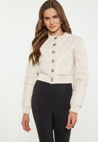Giacca trapuntata color crema con colletto rotondo, dettagli con bottoni dorati e polsini a coste; abbinata a pantaloni neri aderenti.
