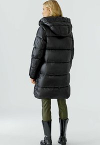 Zwarte pufferjas met een capuchon, met doorgestikte delen. Draagbaar over olijfgroene broek en zwarte knie-laarzen. Glad, glanzend materiaal.