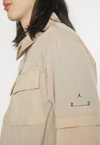 Giacca beige con tessuto strutturato, dotata di tasca sul petto, logo sulla manica e design dal fit rilassato.
