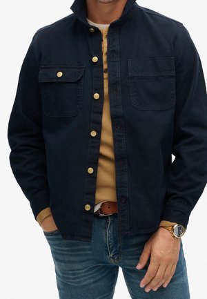 Man wearing dark navy button-up jacket over tan sweater and blue jeans, with hands in pockets and wristwatch on left wrist.