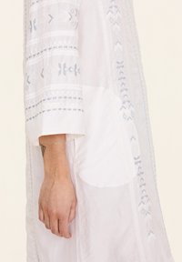 Weißes Kleidungsstück mit hellblauer Stickerei, das geometrische Muster zeigt. Das Design verfügt über eine subtile Textur und kurze Ärmel und präsentiert einen eleganten Schnitt.