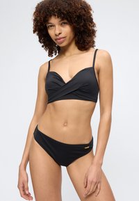 Γυναίκα με σγουρά μαλλιά που φοράει μαύρο μπικίνι με λεπτές τιράντες, στέκεται μπροστά σε απλό φωτεινό φόντο.