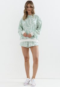 Miętowy komplet z bluzą z kapturem i szortami z białym wzorem fali, wykonany z miękkiego materiału, o luźnym fasonie, z prążkowanymi mankietami i białymi akcentami. W połączeniu z białymi trampkami.