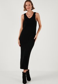 Black ribbed midi dress with a V-neck, sleeveless design. Features a slim silhouette and a frayed hem. Paired with black ankle boots.