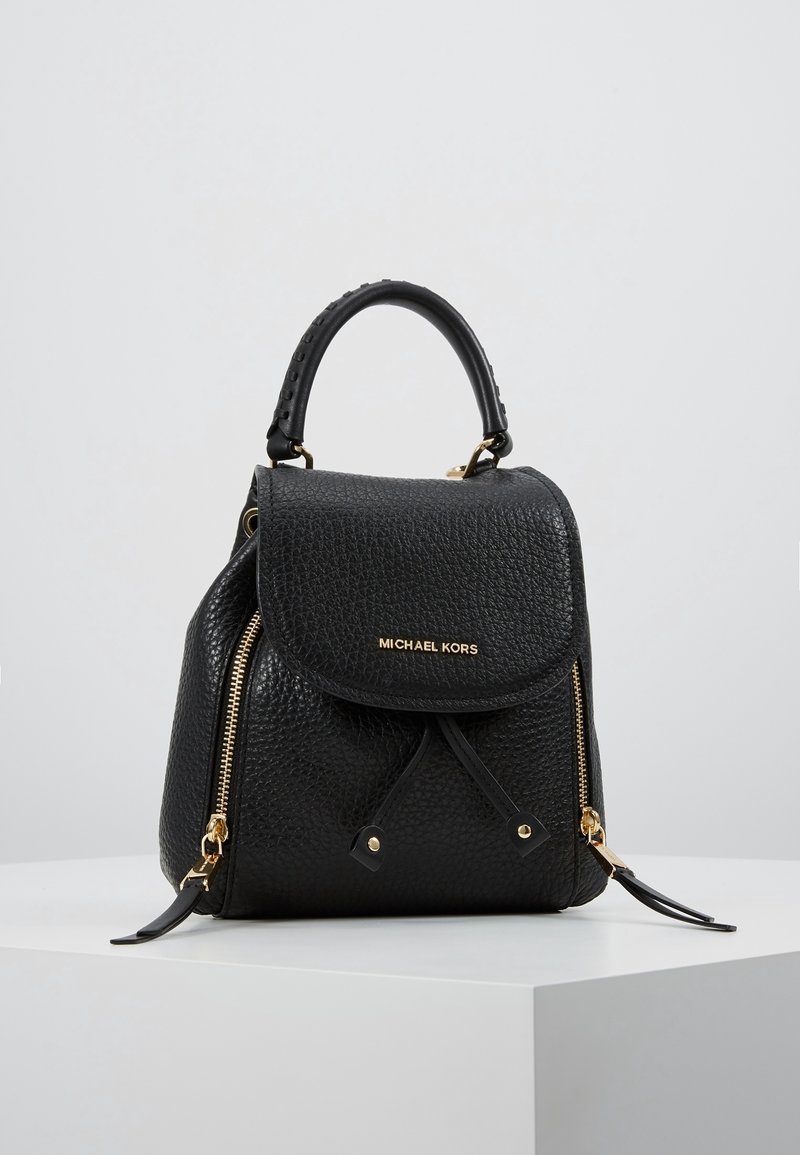 Sac à main Michael Kors en cuir noir texturé avec fermetures éclair dorées, fermeture à rabat et courte anse, présenté sur un socle blanc.