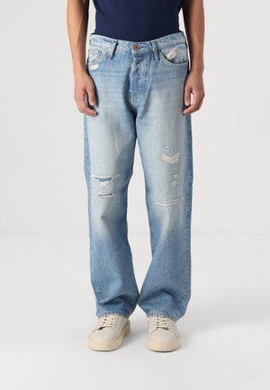 Man draagt losse, lichtblauwe gescheurde spijkerbroek met witte sneakers en een donker shirt, staand tegen een effen achtergrond.