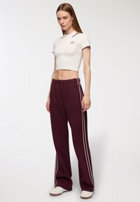 Polo shirt bianco accorciato con rifiniture bordeaux, abbinato a pantaloni della tuta bordeaux con strisce bianche sui lati e vestibilità rilassata. Scarpe bianche.