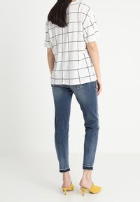 Mujer con camisa blanca de manga corta con un patrón de cuadrícula negra, jeans ajustados azules y mules de tacón amarillo, de pie contra un fondo blanco.