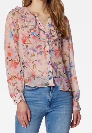 Femme portant un chemisier floral transparent à manches longues avec col froufroutant et boutonnage devant, associé à un jean bleu clair.