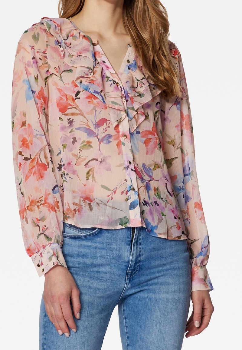 Femme portant un chemisier floral transparent à manches longues avec col froufroutant et boutonnage devant, associé à un jean bleu clair.