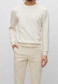 Beige långärmad bomullströja med rund halsringning och en liten logotypmärkt patch på bröstet. Matchas med ljusbeiga byxor i samma färg.