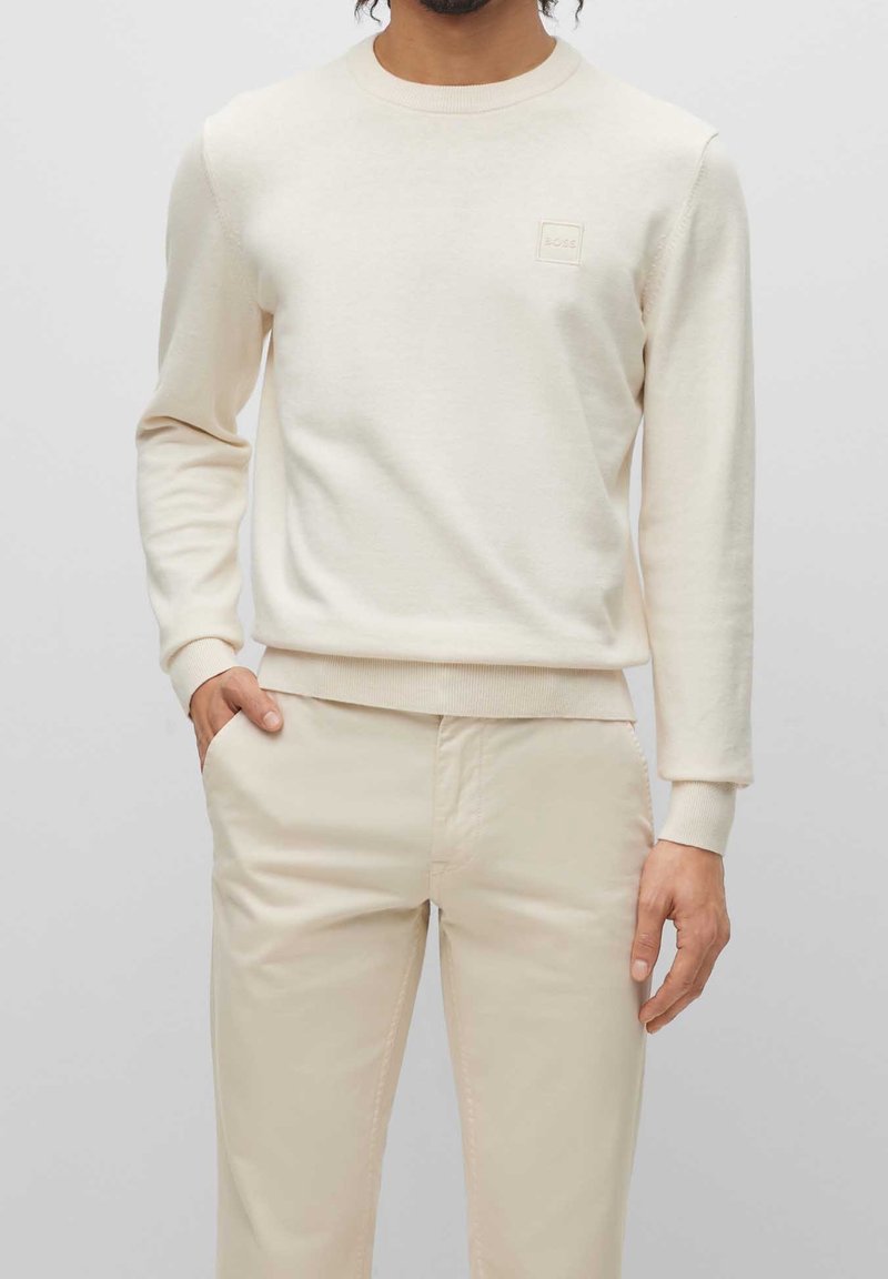 Beige långärmad bomullströja med rund halsringning och en liten logotypmärkt patch på bröstet. Matchas med ljusbeiga byxor i samma färg.