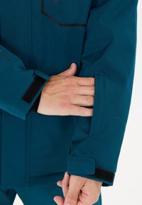 Person steckt die Finger in die Seitentasche einer türkisfarbenen Jacke mit Klettverschluss am Handgelenk vor einem weißen Hintergrund.