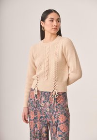 Suéter beige de punto con patrones trenzados, cuenta con lazos laterales, combinado con pantalones estampados con flores en tonos de rosa, verde y naranja.