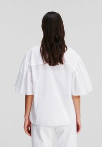 Top bianco a maniche corte con tessuto texturizzato, un motivo delicato e una vestibilità ampia, caratterizzato da maniche a sbuffo e un orlo posteriore arrotondato.