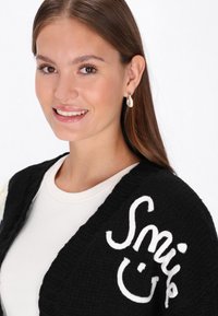 Juodas megztinis su užrašytu baltu "Smile" tekstu. Dėvimas ant šviesaus marškinėlių, su tekstūruotu audiniu ir V formos išpjova.