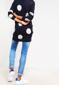 Pull bleu marine à gros pois blancs, manches longues. Associé à un jean bleu clair et des baskets blanches avec des accents bleus.