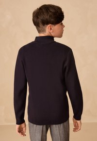 Tumesinine kampsun kõrge kraega ja pikkade varrukatega. Kangas näib pehme ja tekstuuriga, kantud ruuduliste pükstega.