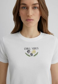 White cotton t-shirt featuring a printed design with the text "CHILL VIBES," colorful flowers, and seashell graphics. Short sleeves and standard fit.