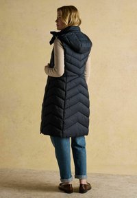 Sieviete ģērbusies garā melnā pūkainā kapuces vestē virs bēša džempera un piegrieztām zilām džinsām, stāv uz strukturēta grīdas pret tukšu sienu.