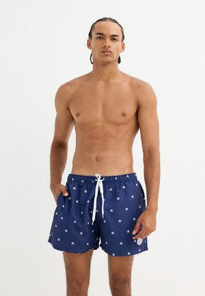 Hombre sin camiseta con cabello trenzado, vistiendo pantalones cortos de baño azul marino con patrones blancos de pulpos y cordón blanco, de pie contra un fondo liso.