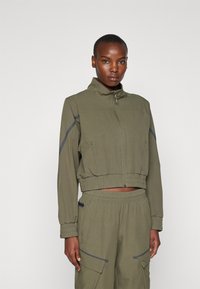 Olivenfarget cropped jakke med høy krage, glidelås foran og elastisk hem. Har to sidelommer og grå aksentstriper på ermene.