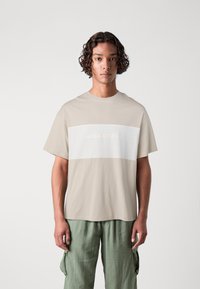 Tamnokrem majica s kratkim rukavima s horizontalnom bijelom trakicom, koja sadrži utisnuti tekst "JACK&JONES". Okrugli izrez.