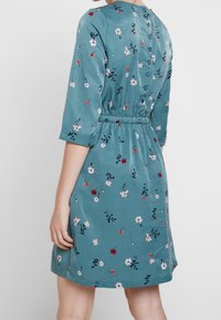 Teal klänning med blommönster, trekvartslånga ärmar, samlad midja och knappstängning bak. Mjuk tyg med en slät textur.