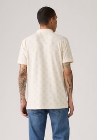 Hellbeige Poloshirt mit kurzen Ärmeln, das ein dezentes eingeprägtes Muster, einen klassischen Kragen und eine lockere Passform aufweist, kombiniert mit blauen Jeans.