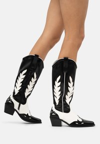 Botas altas en negro y blanco, hechas de material brillante, con patrones de hojas blancas, puntera afilada y un tacón corto y apilado.