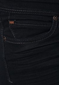 Gros plan sur la poche d'un pantalon en jean noir avec des coutures visibles, des rivets en cuivre et une petite étiquette en cuivre près du coin supérieur de la poche.