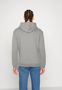 Persona con cabello trenzado vista desde atrás, vistiendo una sudadera con capucha gris y jeans azules, contra un fondo liso.