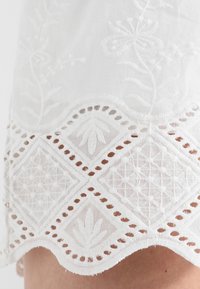 Bíle vyšívaná látka se květinovými a geometrickými vzory. Zvlněný okraj s výřezovými detaily a akcenty poblíž lemu.