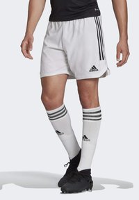 Witte sportshorts met zwart Adidas-logo, gecombineerd met knielange witte sokken met zwarte strepen. Zwarte voetbalschoenen maken de look compleet.