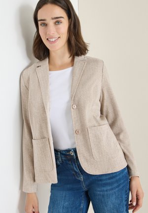 Blazer en chevrons beige avec deux poches, fermeture à un bouton et revers crantés, associé à un haut blanc et un jean bleu.