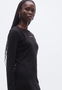 Juoda ilgas rankovėmis aptempta suknelė su apvalia iškirpte, krūtinės srityje subtilus logotipas. Lygus audinys ir elegantiška siluetas.