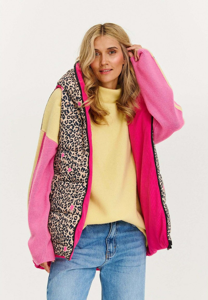 Barevná vesta s panely v růžovém a leopardím vzoru, texturovaná látka, zapínání na zip a oversize střih, doplněná žlutým pleteným svetrem.