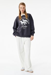 Donkergrijze oversized sweatshirt met een witte paardenafbeelding en tekst, gecombineerd met lichtbeige wijde broek en grijze schoenen.
